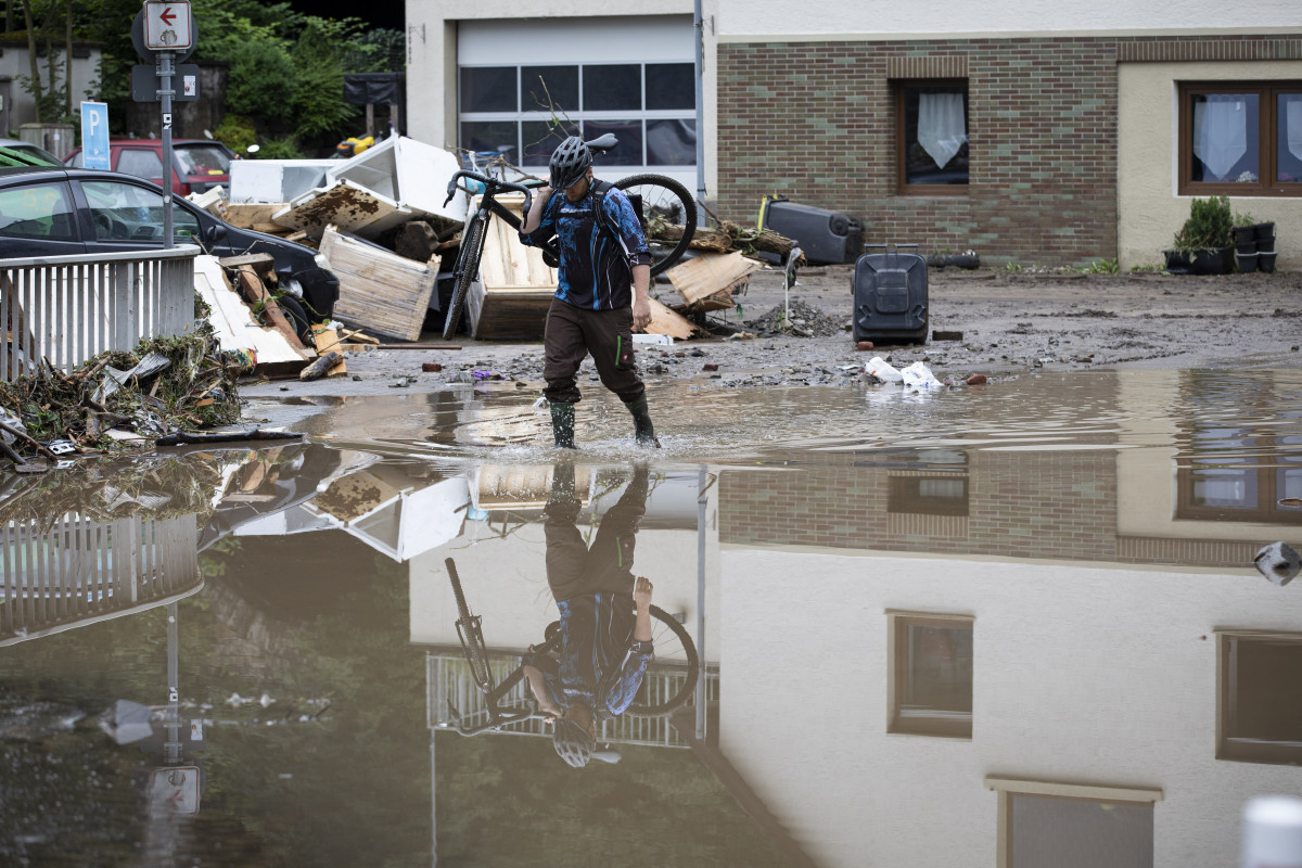 Photo shows man with bicycle over shoulder wading through flooding in city of Altena in Germany 2021. Photo: Land NRW / Ralph Sondermann. 